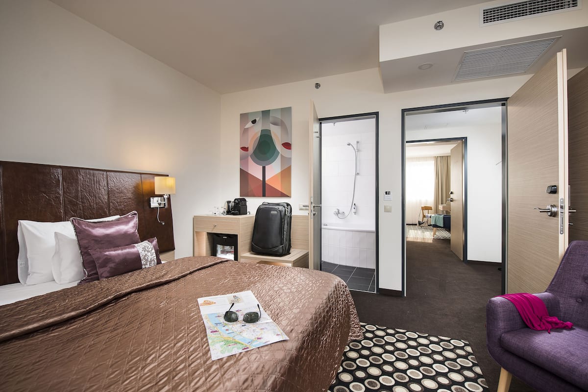 A hotel room with a bed, a desk, a chair, a bathroom, and a hallway.