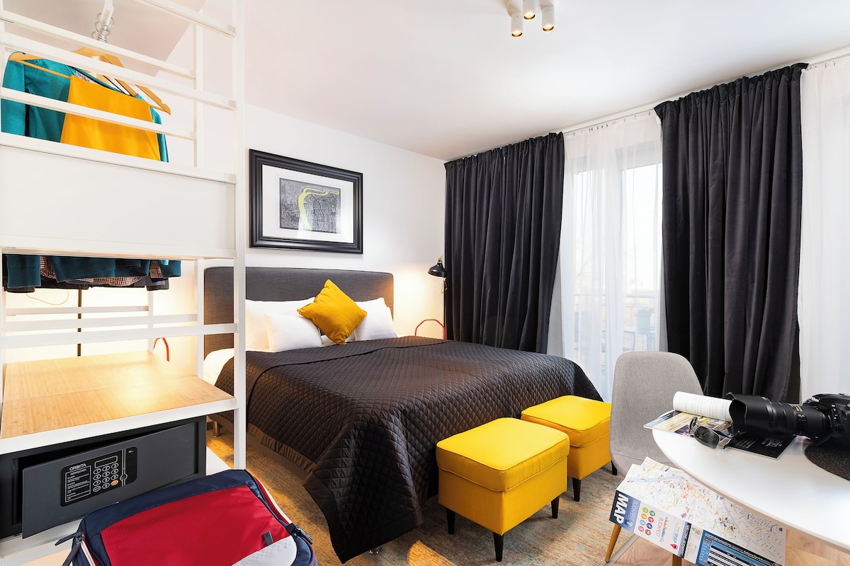 A hotel room with a bed, a desk with a camera, a chair, a desk lamp, a bookshelf, and a window with curtains.