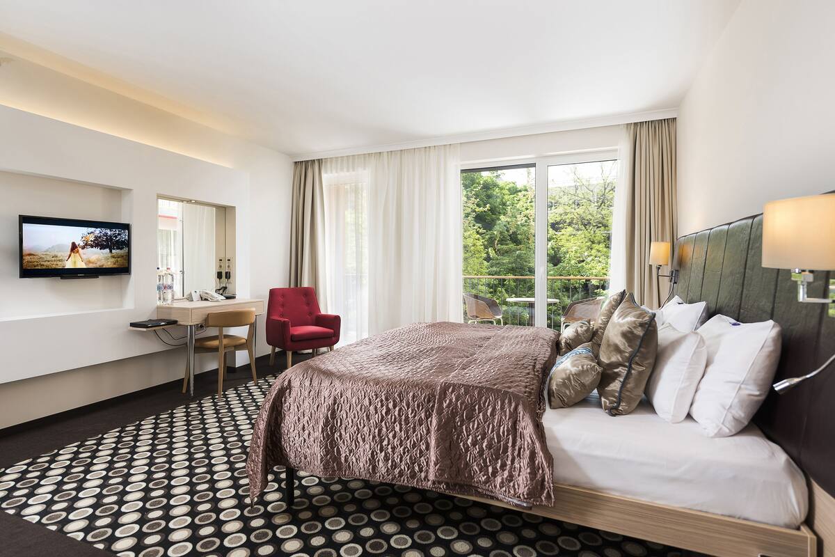 A hotel room with a bed, a desk with a chair, a television, and a large window with curtains.