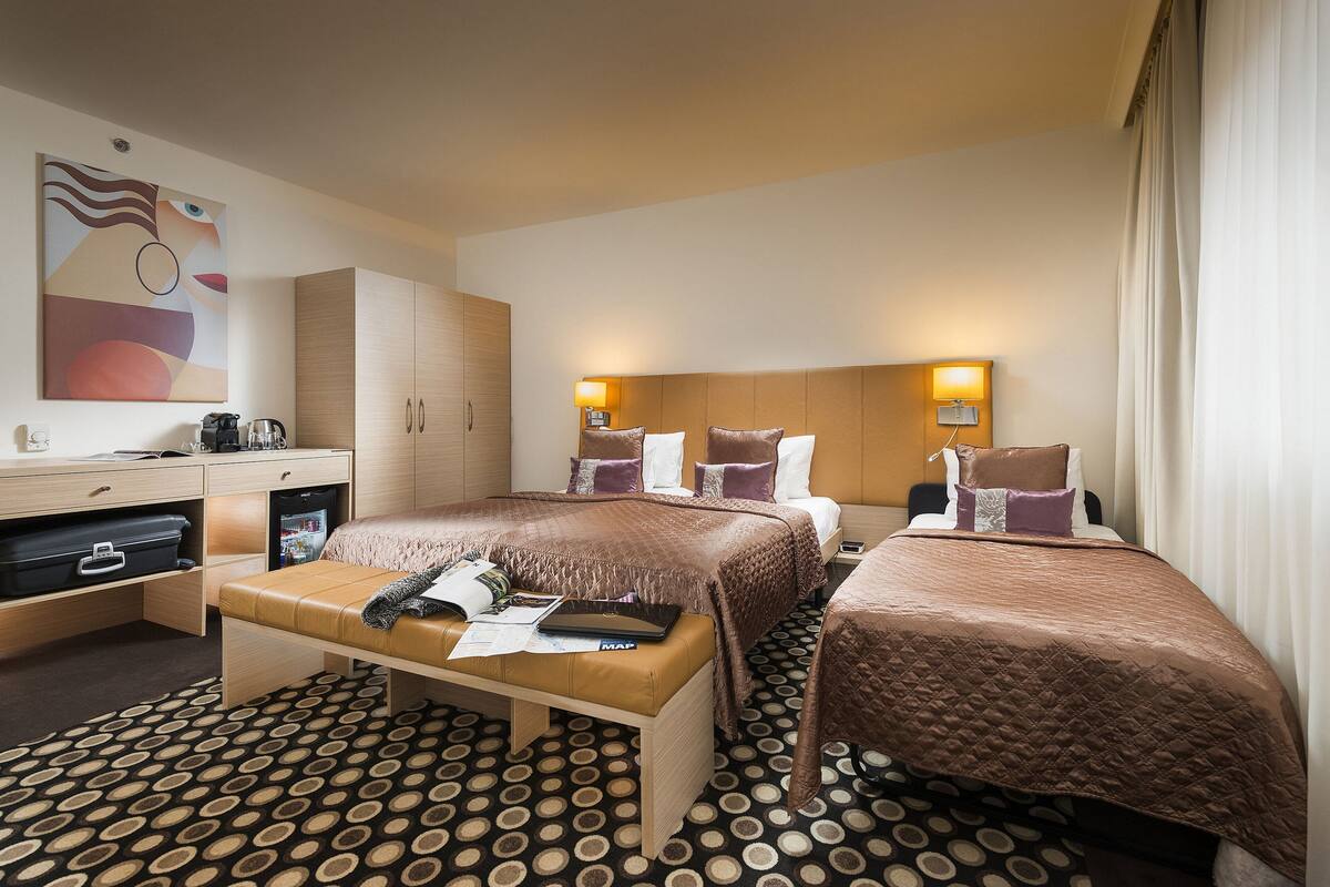 A hotel room with two beds, a desk with a computer, a small fridge, and a patterned carpet.