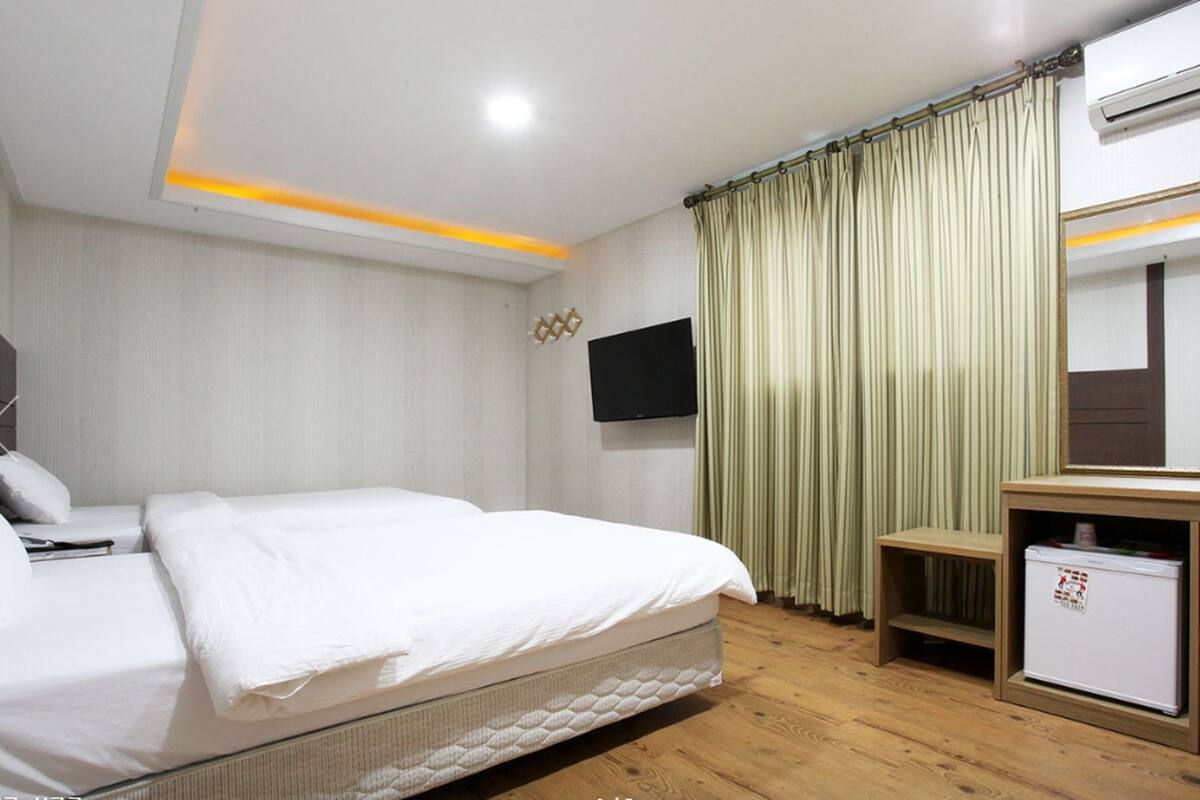 Habitación de hotel con cama, televisión fijada a la pared, un mueble pequeño y ventana con cortinas.