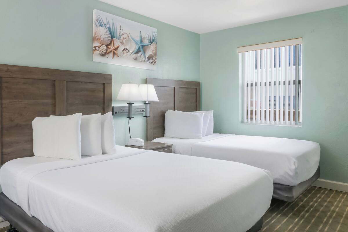A hotel room with two beds, a wooden headboard, a painting of seashells, a nightstand with a lamp, and a window with blinds.