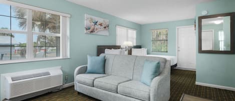 A room with a grey sofa, a bed, a window, and a wall-mounted mirror.