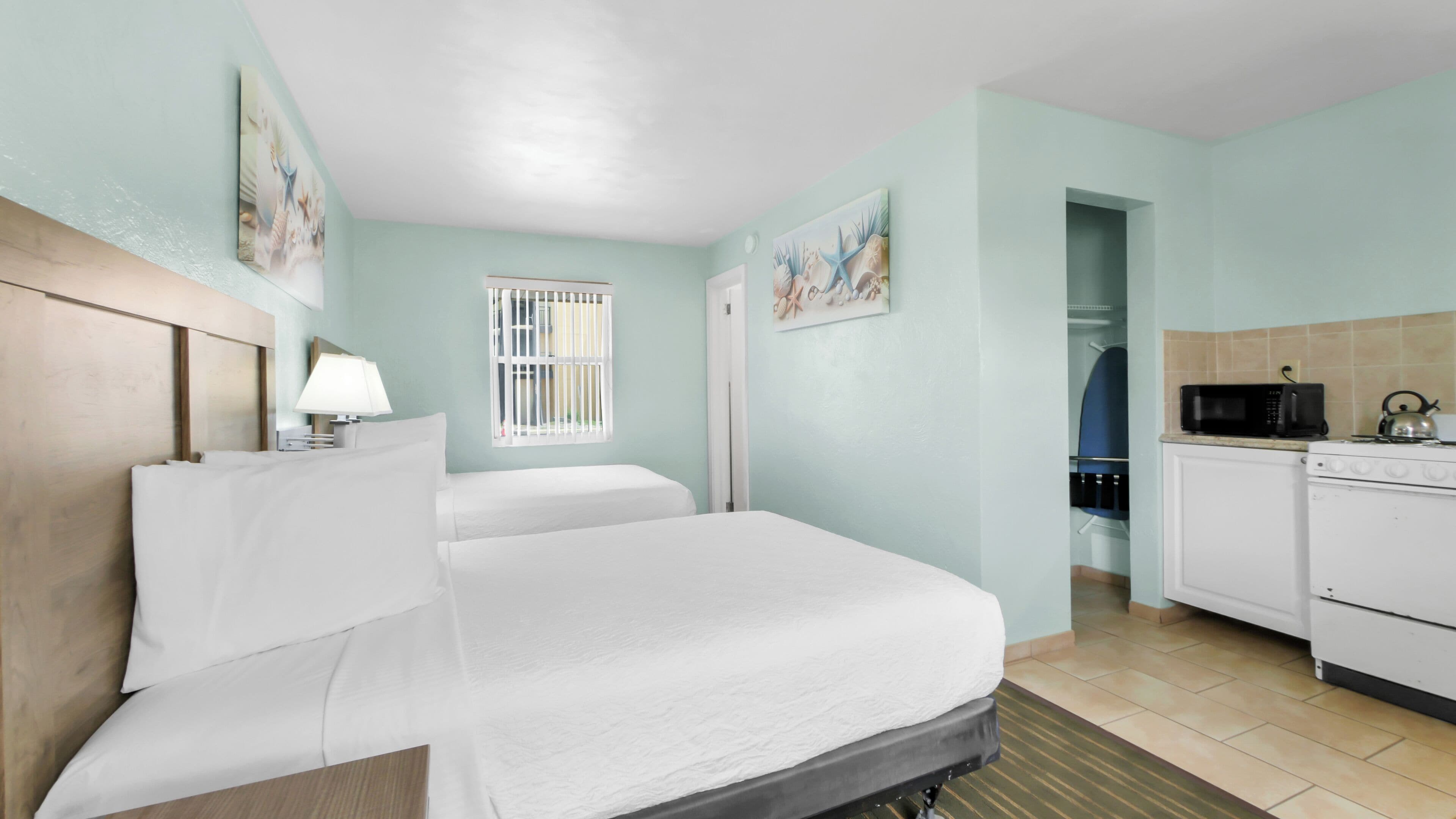 A hotel room with a bed, a bedside table with a lamp, a window with bars, a microwave, a kettle, and a small kitchenette.