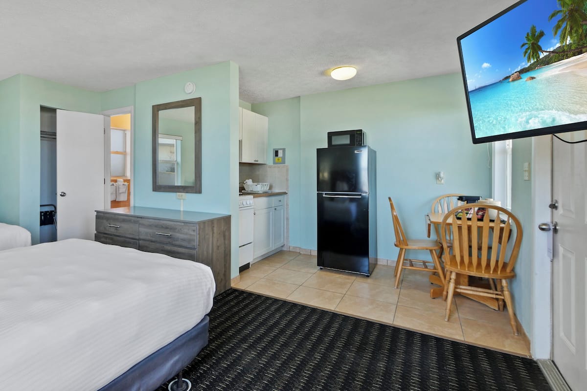 A hotel room with a bed, a desk with a chair, a refrigerator, a microwave, and a television displaying a beach scene.