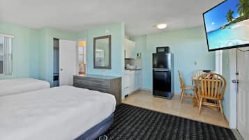 A hotel room with a bed, a desk with a chair, a refrigerator, a microwave, and a television displaying a beach scene.