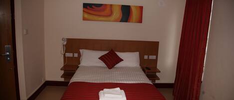A hotel room with a large bed, a red and white striped bedspread, a red pillow, a red curtain, a wall-mounted artwork, and a bedside table with a lamp.
