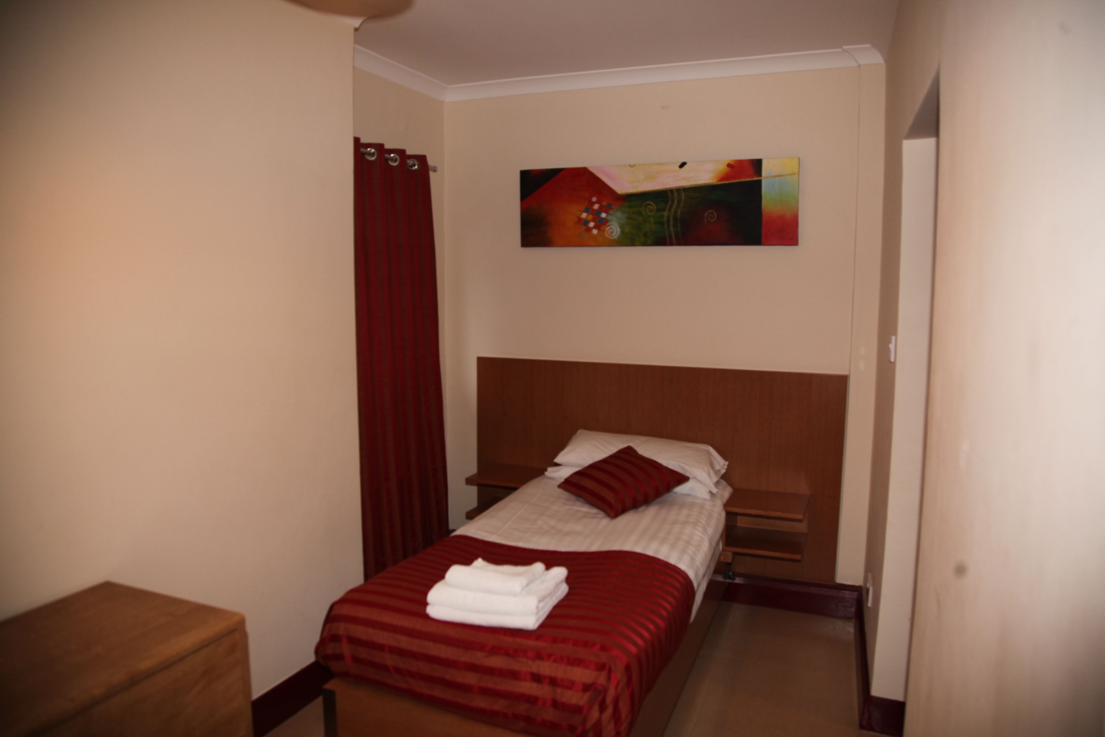 A small, neatly made bed with a red bedspread and pillows, a wooden nightstand, and a painting on the wall.
