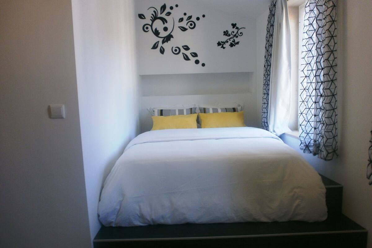 A bedroom with a bed, a window with patterned curtains, and a wall decoration.