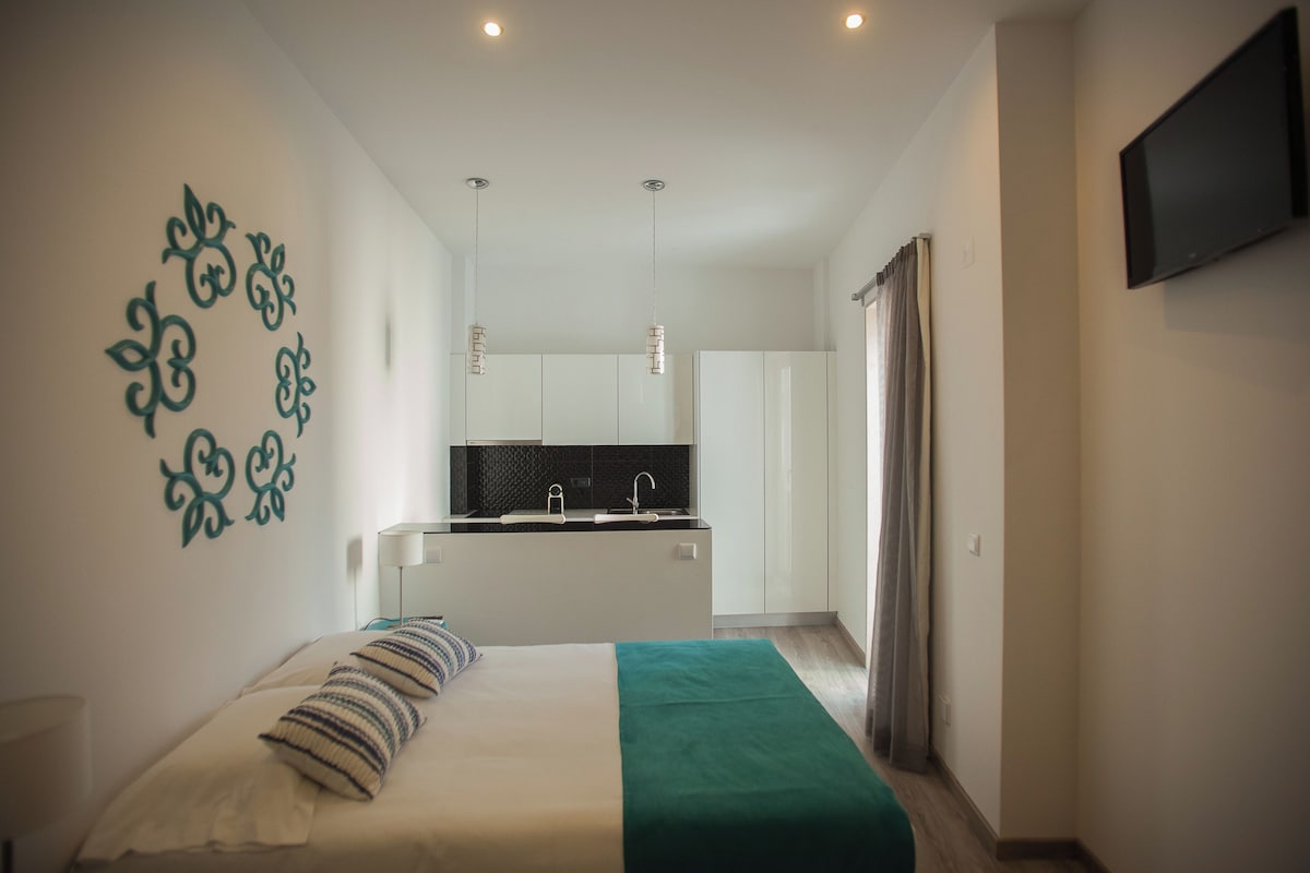 A modern hotel room with a bed, a green throw blanket, a kitchen area with white cabinets, and a flat-screen TV mounted on the wall.