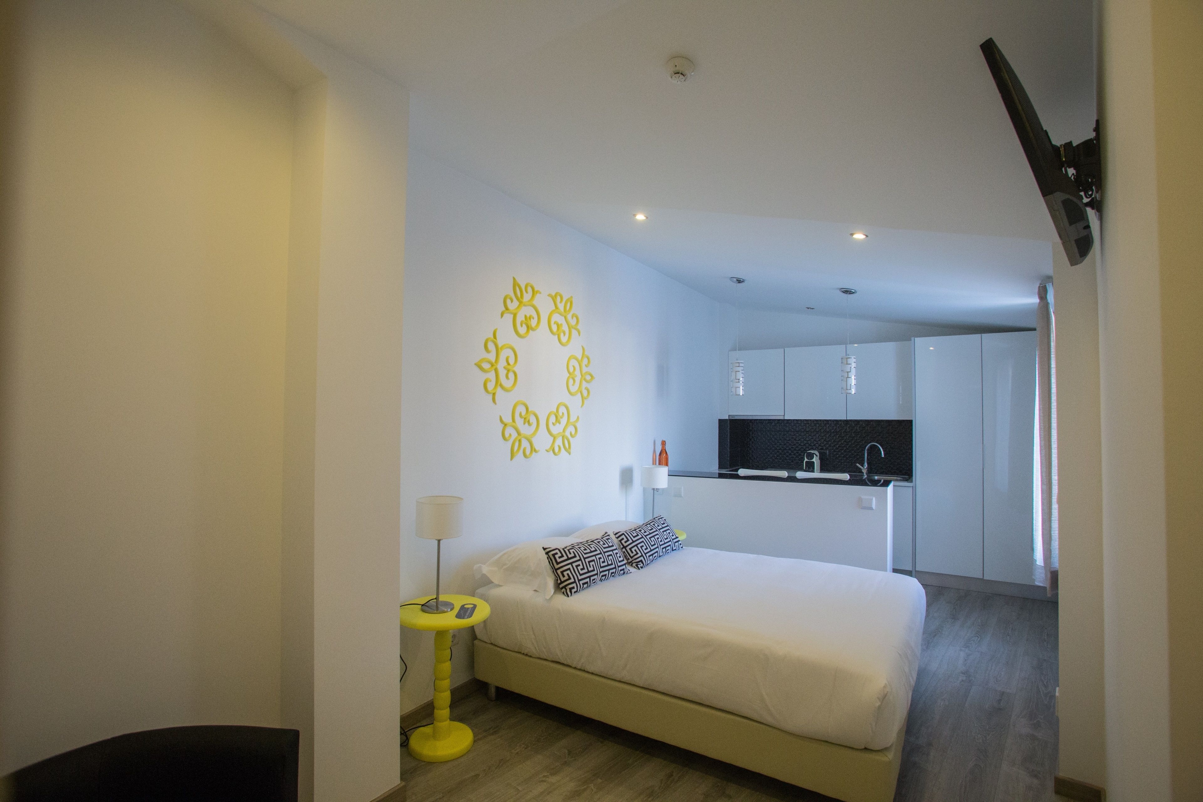 Un dormitorio moderno con cama, mesita de noche y un escritorio pequeño. Se ve una cocina al fondo.