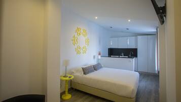 Un dormitorio moderno con cama, mesita de noche y un escritorio pequeño. Se ve una cocina al fondo.