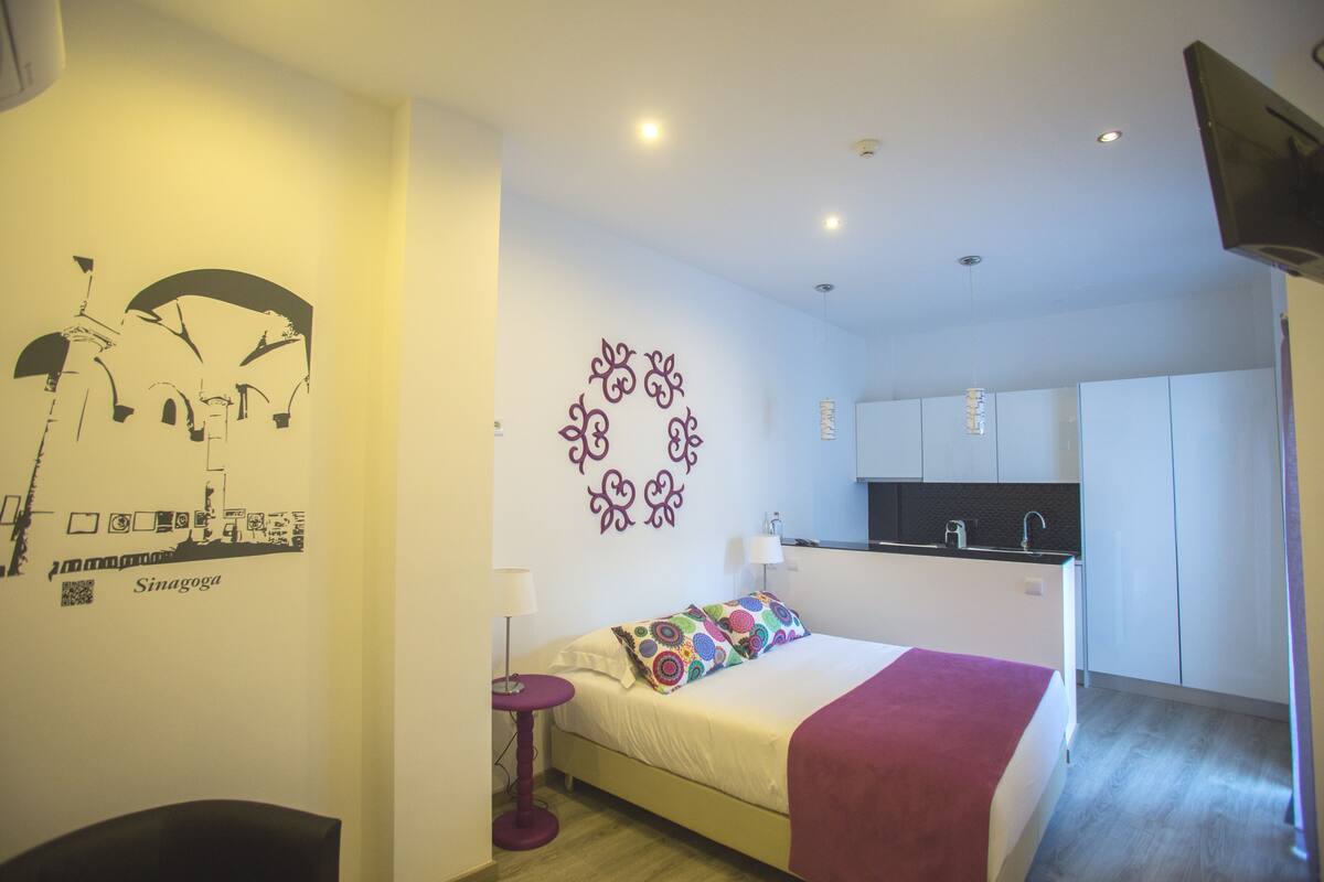 A hotel room with a bed, a small table, a wall decoration, and a kitchenette.
