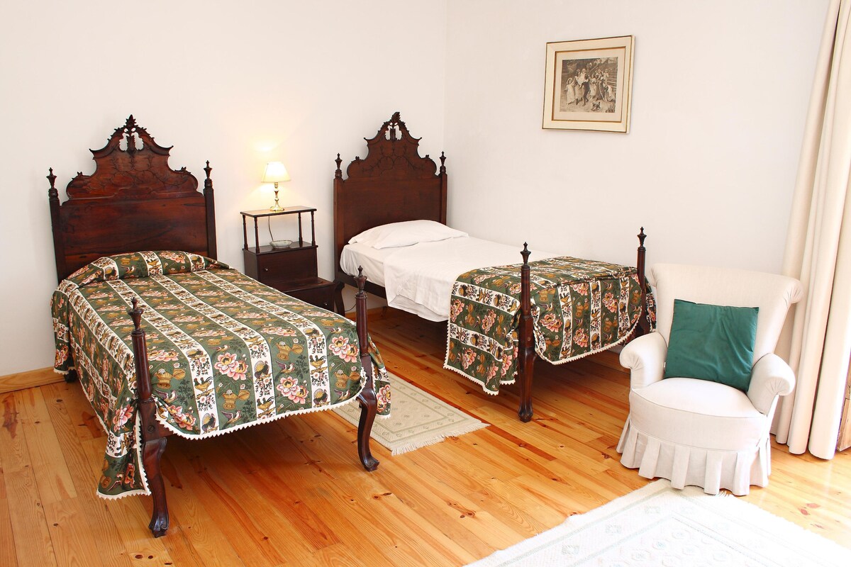 Una habitación con dos camas de madera con edredones florales, un sillón blanco, una mesita y un cuadro enmarcado en la pared.