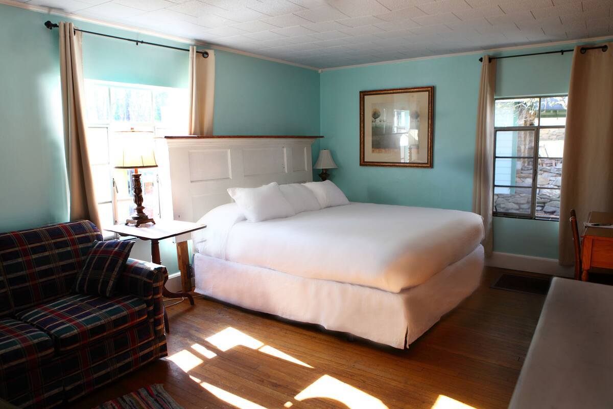 A room with a bed, a plaid sofa, a small table with a lamp, and a window with curtains.
