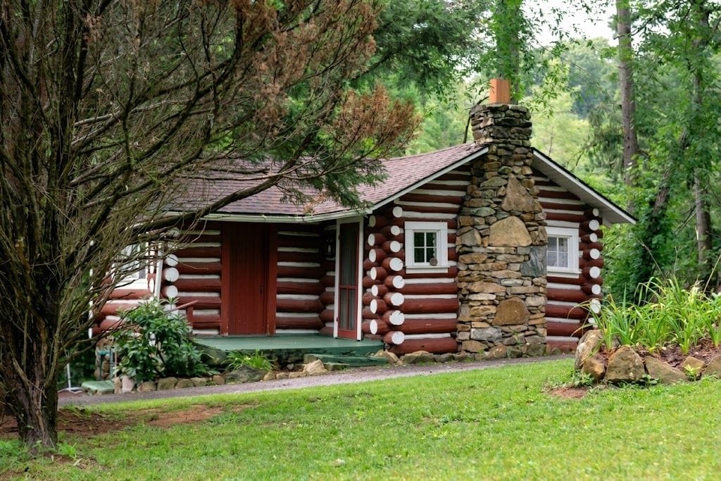 A log cabin with a stone chimney, a porch, and a small window.