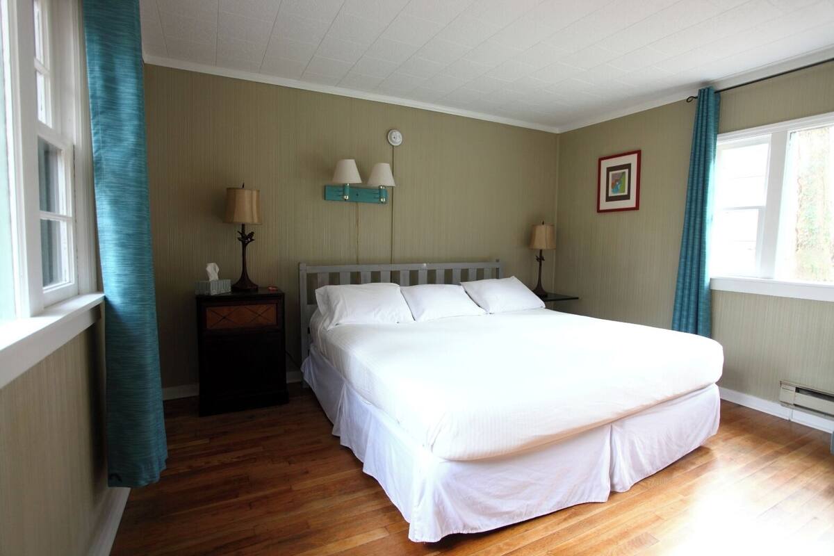 A bedroom with a large bed, wooden flooring, two bedside tables with lamps, a wall clock, and a window with curtains.