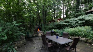 An outdoor patio with a stone fire pit, surrounded by lush greenery and a rustic wooden table with chairs. - The Pines Cottages (Asheville)
