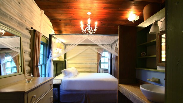 A bedroom with a canopy bed, wooden flooring, a dresser, a bedside table, and a large mirror. - The Pines Cottages (Asheville)