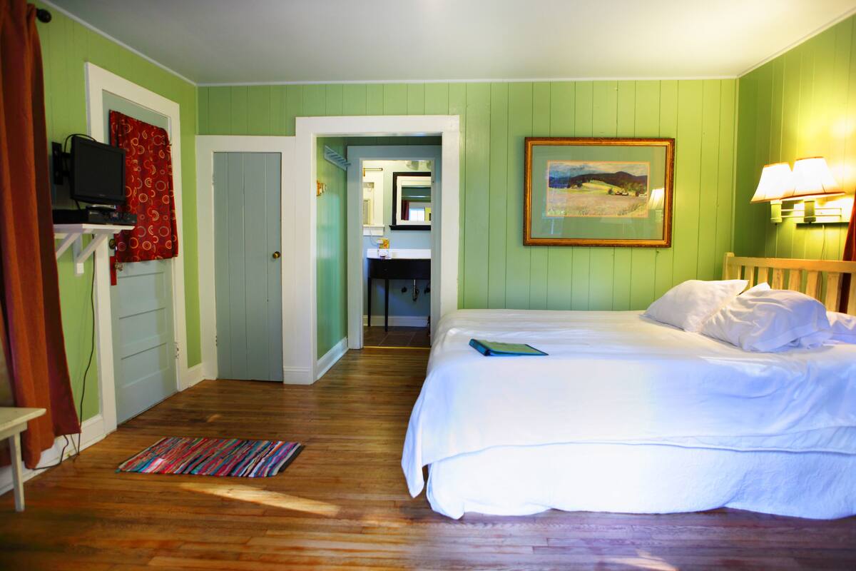 A bedroom with a bed, a television on a shelf, a painting on the wall, and a bathroom visible through an open door.
