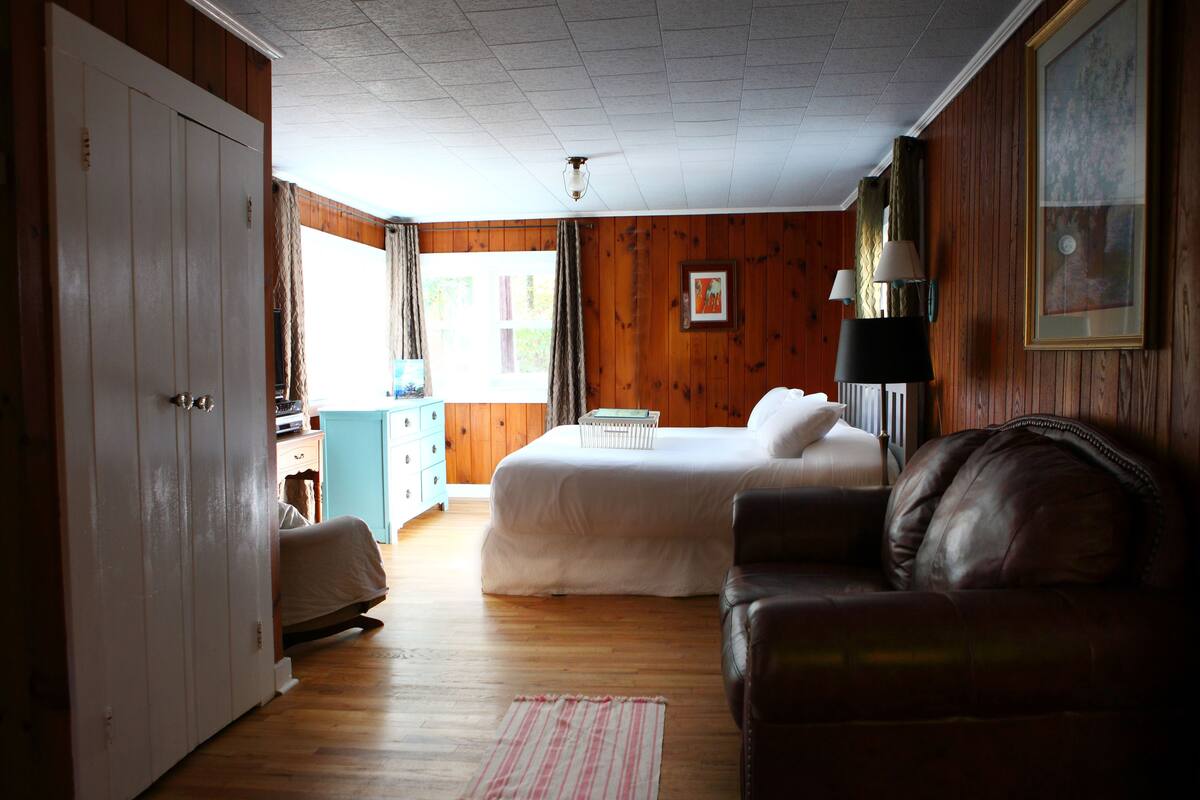 A room with wooden walls, a bed, a desk, a chair, and a lamp.