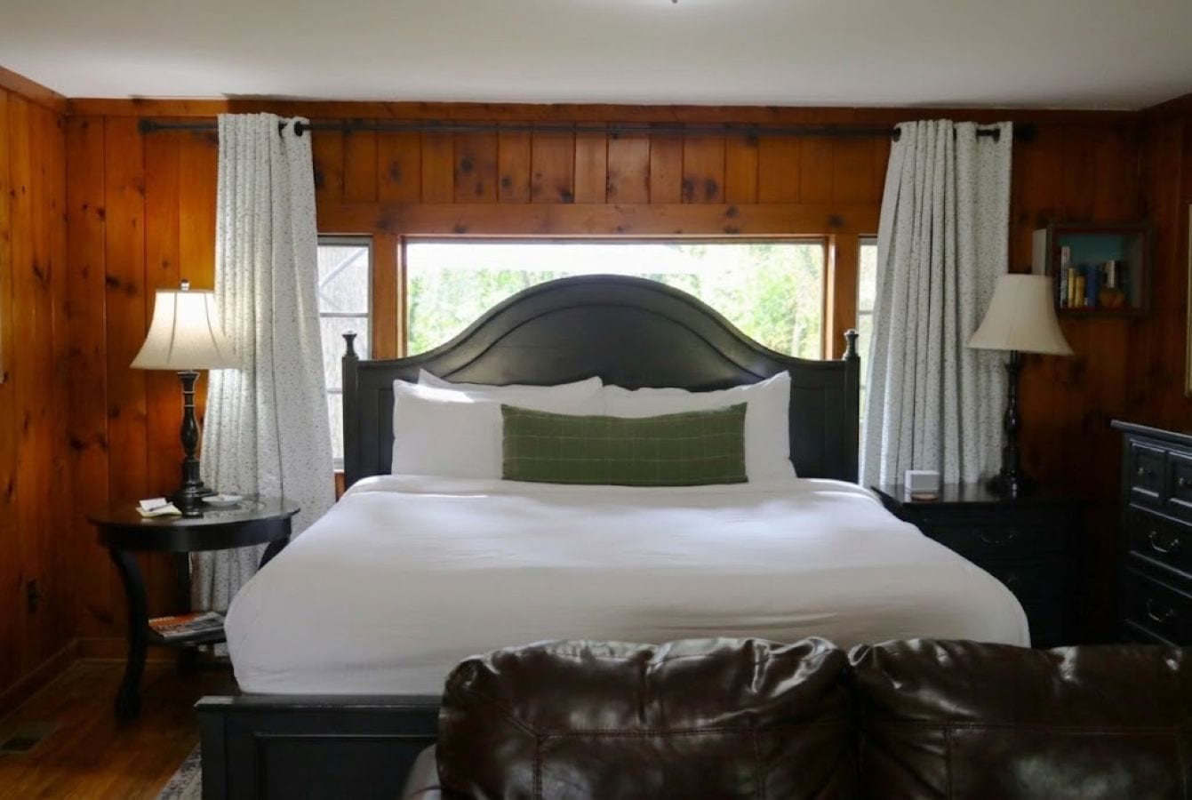 Una habitación con una cama grande, paredes de madera, una ventana con cortinas, una mesita de noche con lámpara y un aparador.
