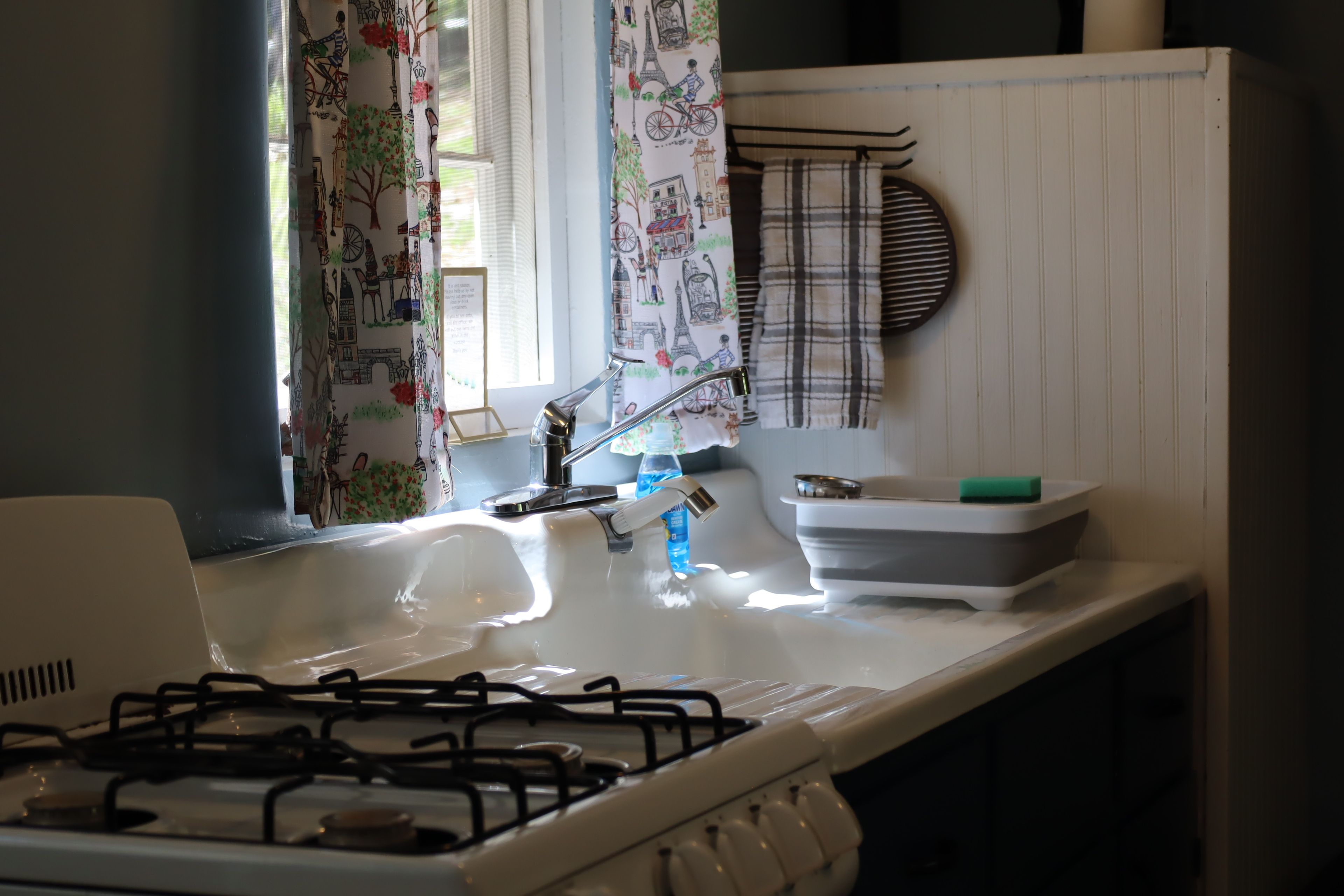 A kitchen with a gas stove, a sink, and a window with decorative curtains.
