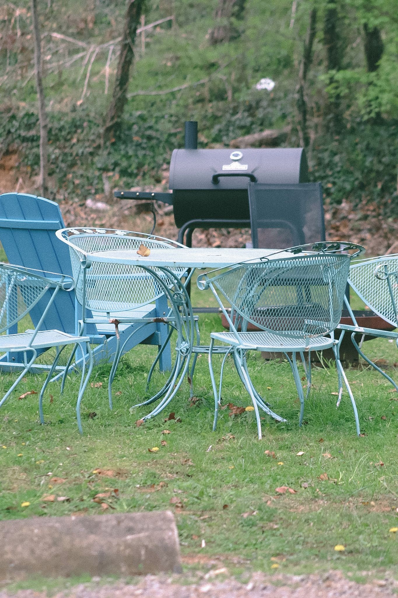 Sillas metálicas y una mesa al aire libre sobre césped, con una parrilla de fondo.