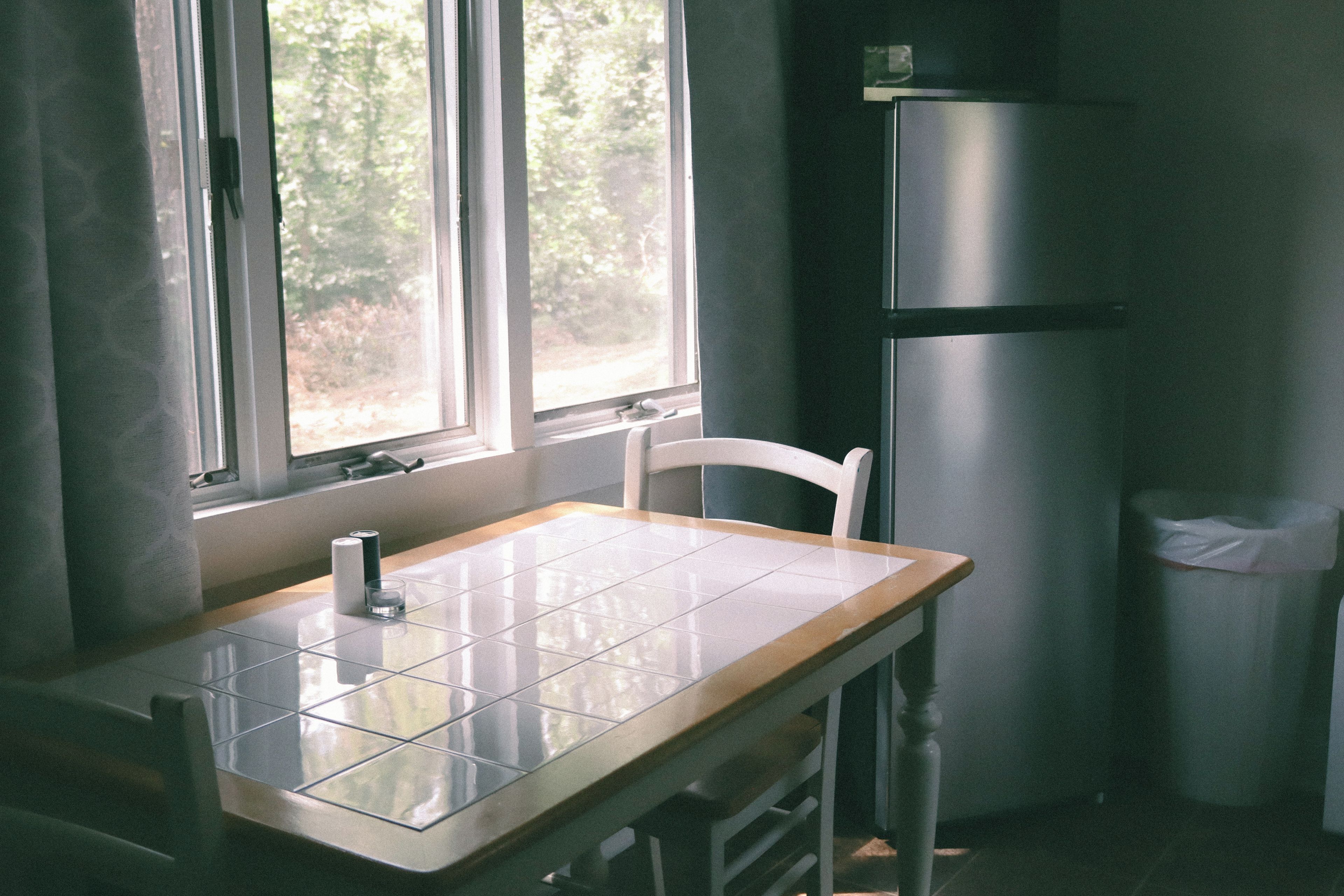 Une cuisine avec un plan de travail carrelé, une chaise, un réfrigérateur et une fenêtre donnant sur des arbres.