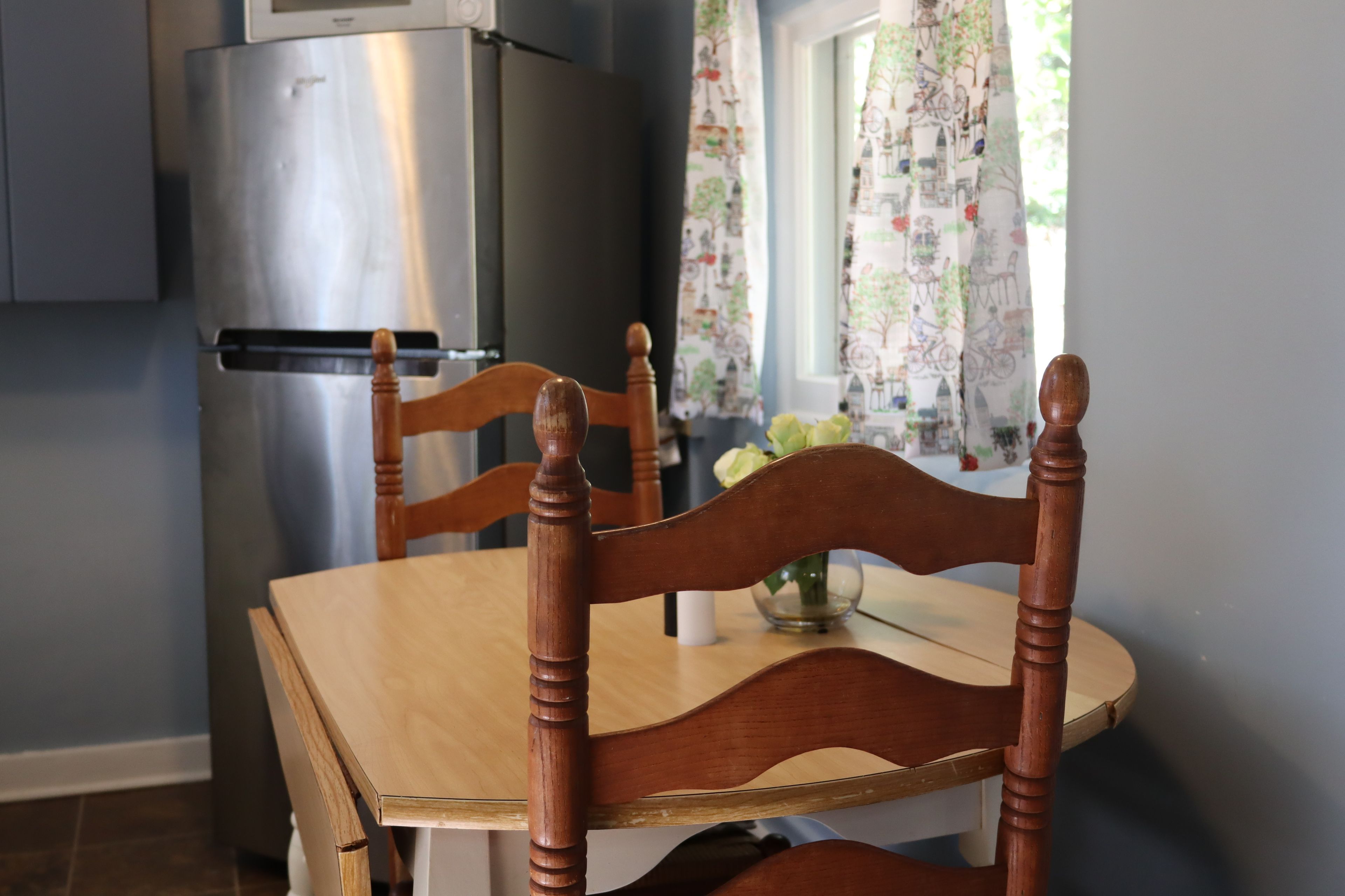 A wooden dining table with chairs, a refrigerator, and a window with curtains.
