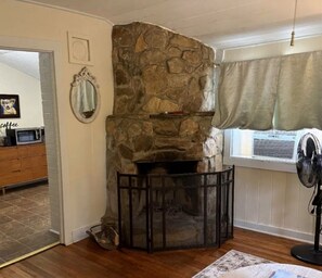 A room with a stone fireplace, a wooden cabinet, a fan, and a window with curtains. - The Pines Cottages (Asheville)