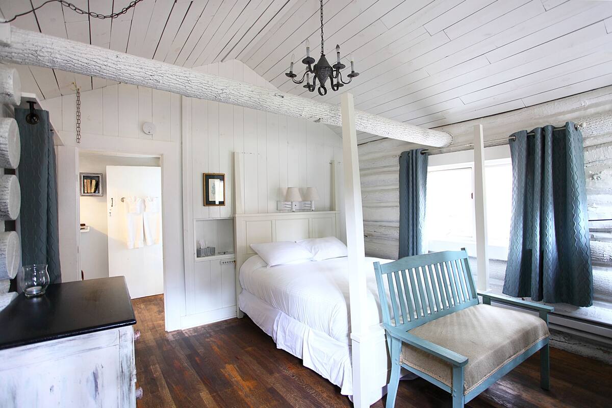 A bedroom with a wooden ceiling, a bed, a chair, a dresser, and a window with curtains.