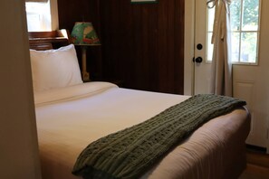 A bed with a white mattress and a green blanket, a lamp with a colorful design, and a door with a curtain. - The Pines Cottages (Asheville)