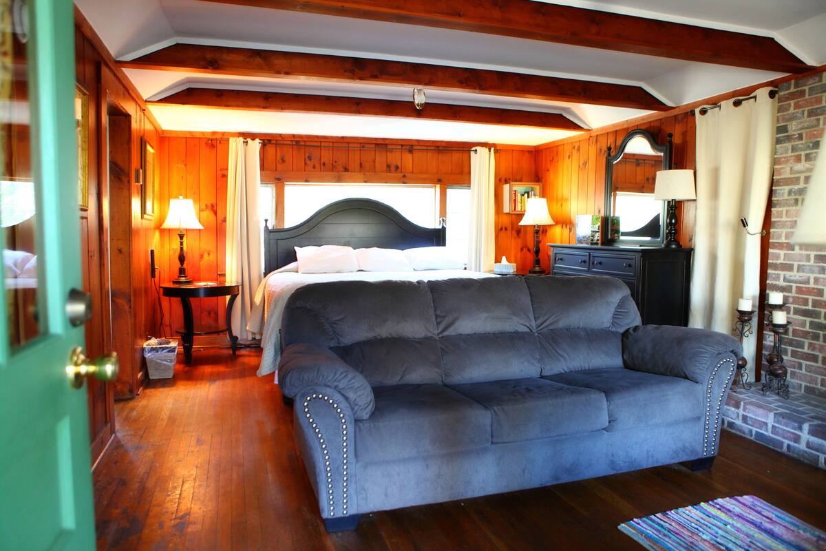 A bedroom with a large bed, a dark gray sofa, wooden walls, and a brick fireplace.