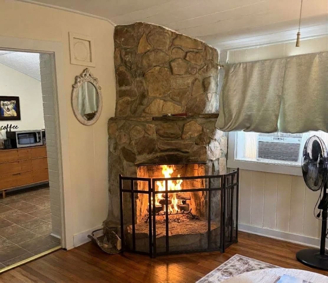 A stone fireplace with a fire, a wooden cabinet, and a fan in a room with wooden flooring.
