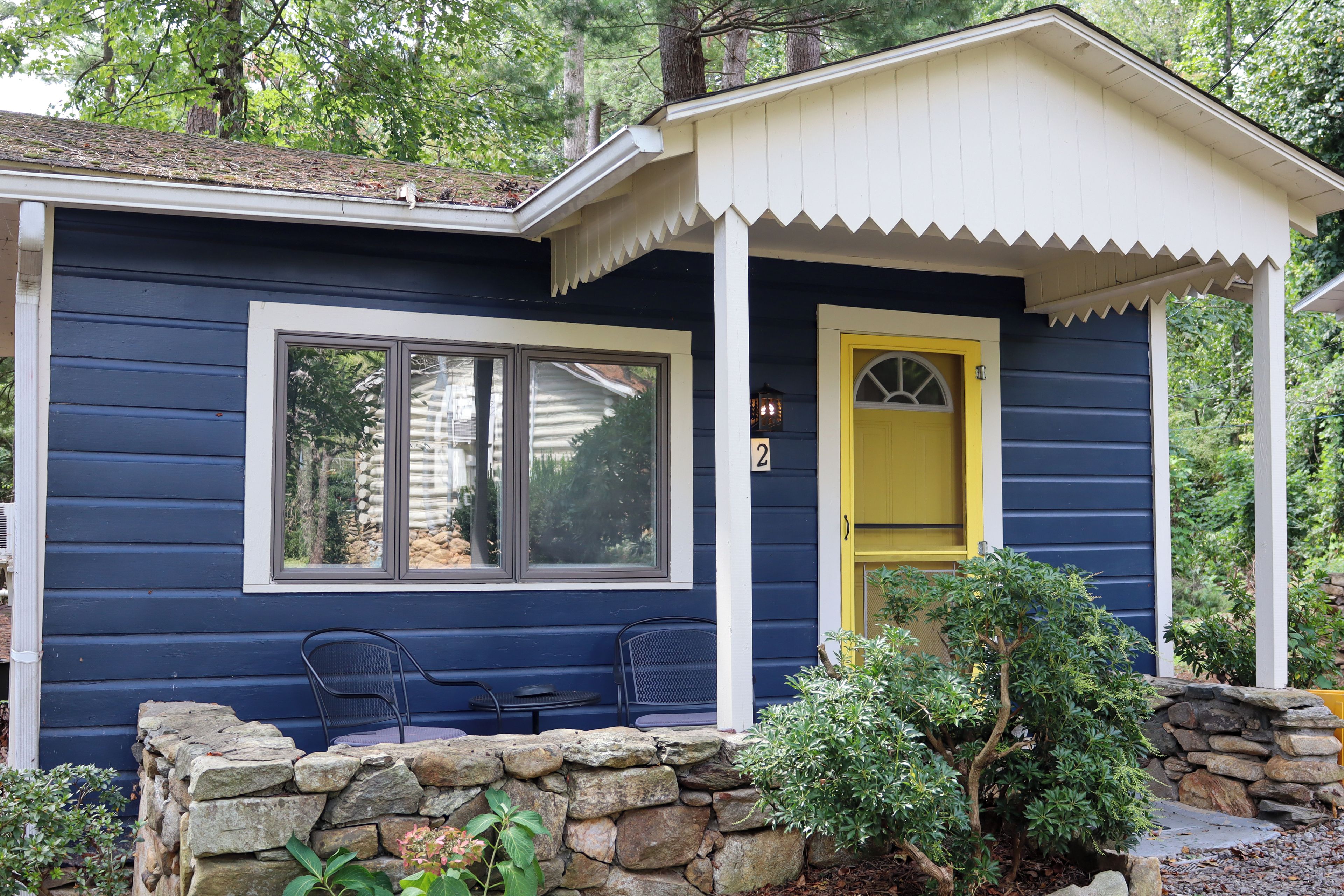 A small, single-story building with a dark blue exterior, a yellow door, and a stone wall in front. There are two windows and a small porch area with chairs.