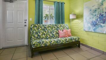 Une pièce avec un mur vert, une porte blanche, un canapé à motifs végétaux, un coussin rose et un tableau.