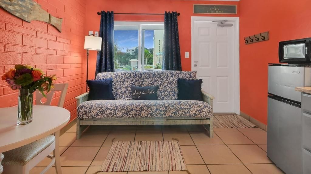 A kitchenette with a microwave and a refrigerator, a sofa with a floral pattern, a small table with a vase of flowers, and a door with a sign.