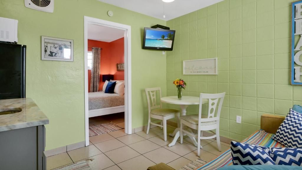 Un espacio habitable compacto con kitchenette, una zona de comedor con dos sillas, una cama con cabecera colorida y un televisor montado en la pared.