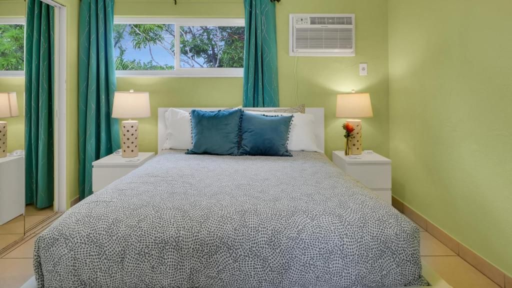 A bedroom with a bed, two bedside tables, a window with curtains, and a wall-mounted air conditioner.