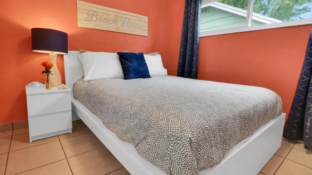 A bed with a patterned bedspread, a bedside lamp, a vase with a flower, and a sign on the wall.