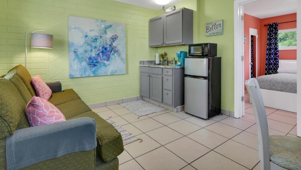 A compact living space with a kitchenette, a sofa with patterned cushions, a painting on the wall, and a sign that reads 'Better at the beach.'