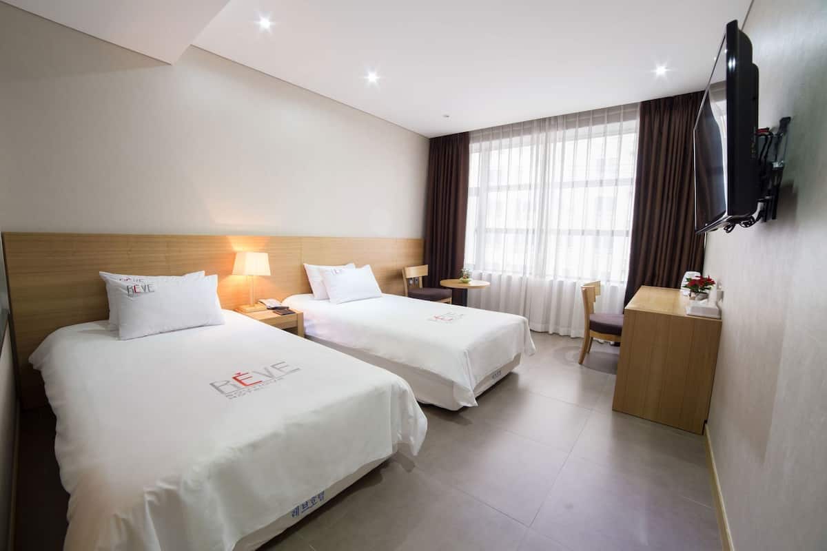 Une chambre d’hôtel avec deux lits, un bureau, une chaise, une télévision et une fenêtre avec des rideaux.