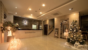 Zona de recepción de un hotel con piso de mármol, mostrador de recepción con la leyenda “INFORMACIÓN”, un árbol de Navidad y una escalera que conduce a los pisos superiores.