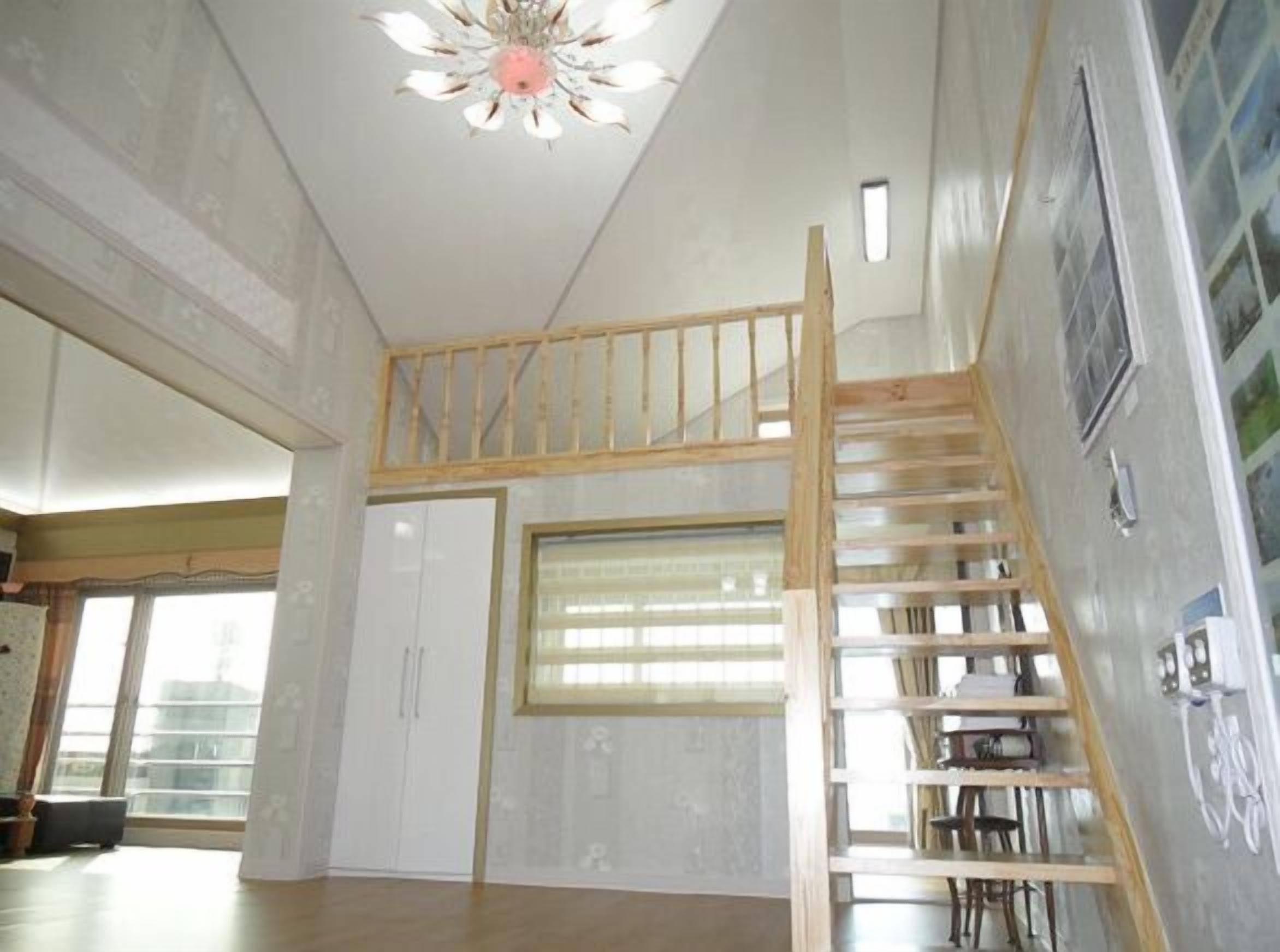 A room with a wooden staircase leading to a loft area, a large window with blinds, and a ceiling fan.