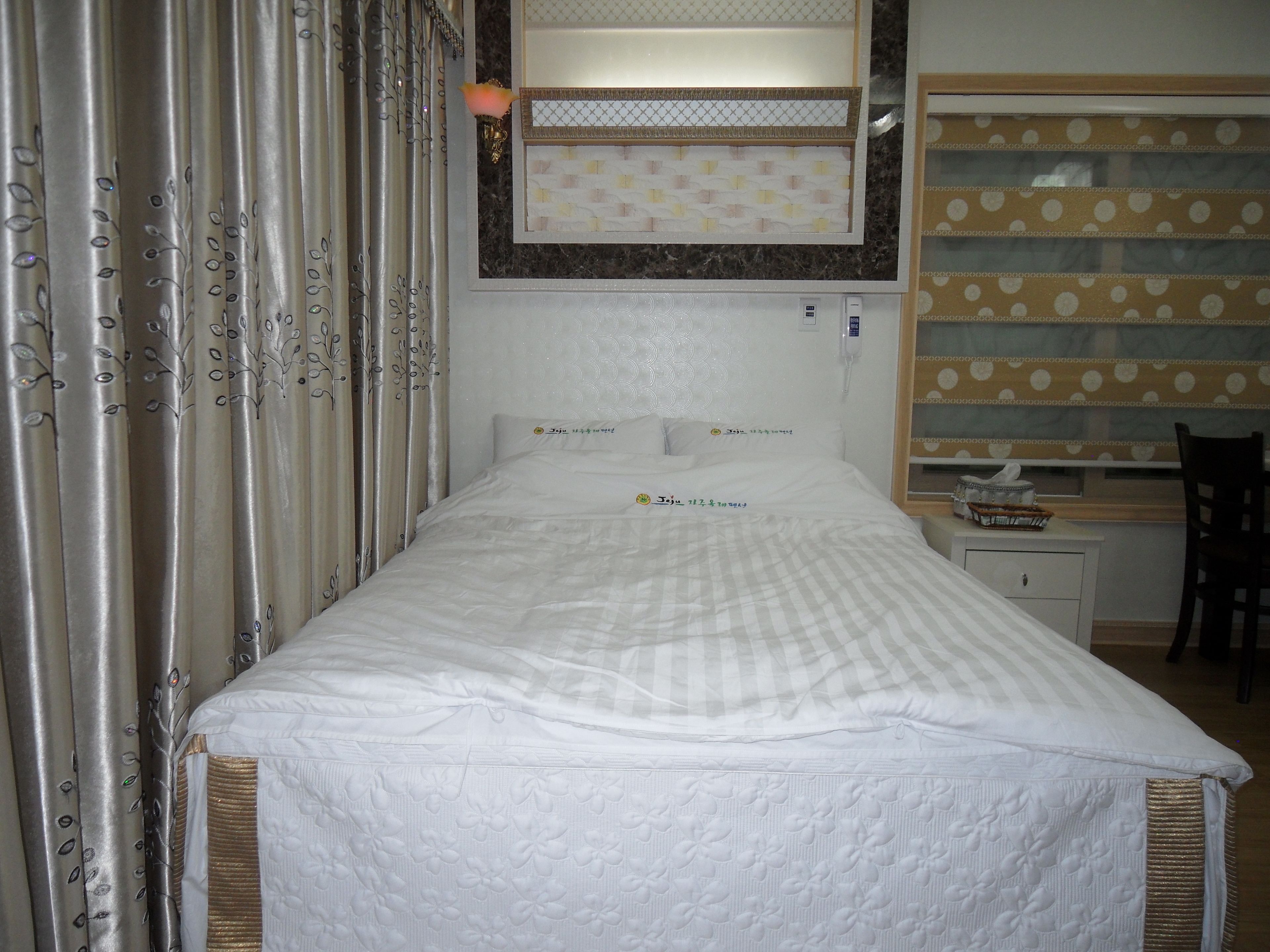 A bedroom with a bed, a desk, and a chair. There is a window with curtains, a wall-mounted phone, and a small bedside table.
