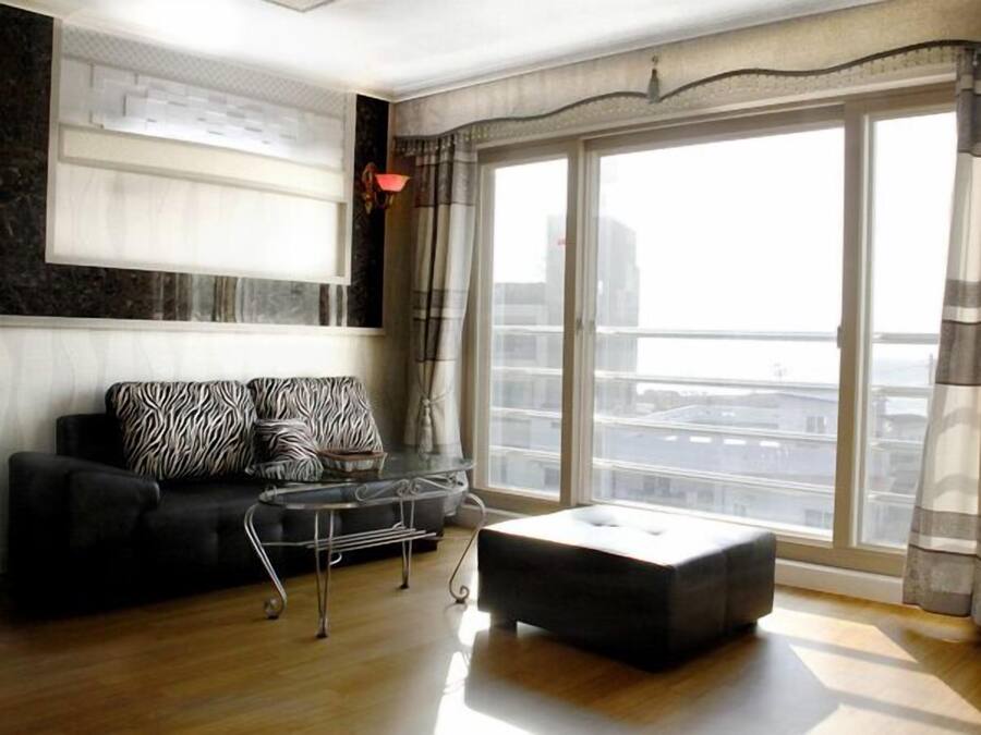 A modern living room with a black sofa, a glass coffee table, and a large window with curtains.