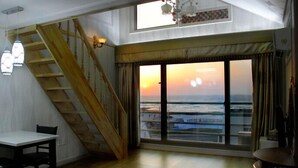 A room with a wooden staircase, a dining table, and a large window with a view of the ocean at sunset. - Jaju Olle Pension (Jeju City)