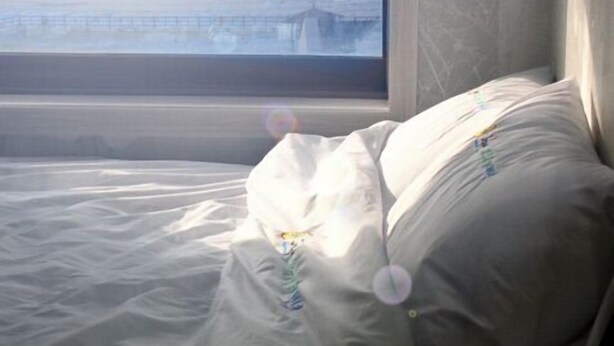A bed with a white comforter and pillows, a window with a view of a beach and ocean, and a gray headboard. - Jaju Olle Pension (Jeju City)