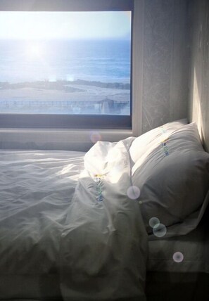 A bed with a white comforter and pillows, a window with a view of a beach and ocean, and a gray headboard. - Jaju Olle Pension (Jeju City)
